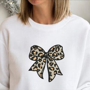 May include: White sweatshirt featuring a leopard print bow design. The bow has black outlines and tan and black spots. The sweatshirt is a casual style, suitable for everyday wear. The design is centered on the chest.