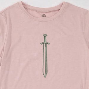 May include: A light pink t-shirt featuring a detailed illustration of a sword. The sword is depicted in a vertical orientation, with a long, slender blade and a cross-shaped hilt. The shirt has short sleeves and a crew neck.