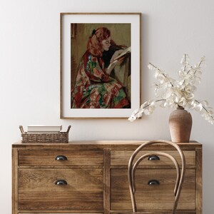 May include: A framed painting of a woman with red hair sitting in a chair and reading a book. She is wearing a colorful patterned dress.