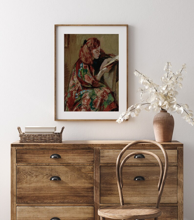 May include: A framed painting of a woman with red hair sitting in a chair and reading a book. She is wearing a colorful patterned dress.