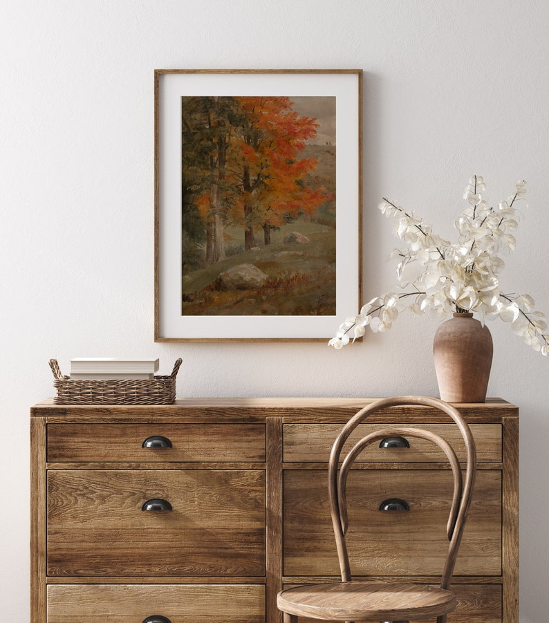Puede incluir: Una pintura enmarcada que representa una escena de bosque con &aacute;rboles en varios tonos de verde y naranja. Los &aacute;rboles est&aacute;n en primer plano de la pintura, con una ladera en el fondo. La pintura est&aacute; colgada en una pared blanca sobre una c&oacute;moda de madera.