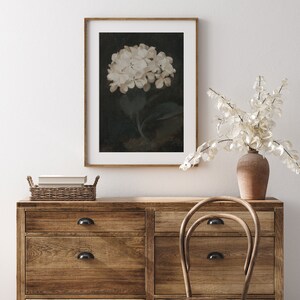 May include: A framed floral print featuring a white hydrangea against a dark background. The artwork is displayed on a wooden dresser with a wicker basket and a vase of dried flowers. A wooden chair sits nearby, creating a cozy, rustic aesthetic.