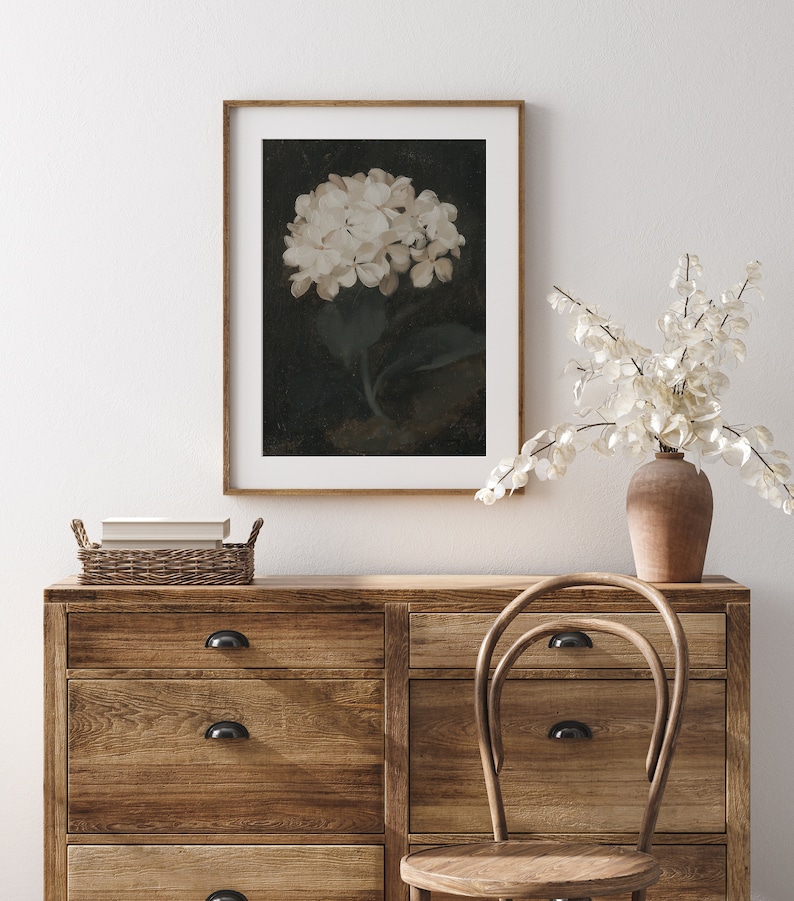 May include: A framed floral print featuring a white hydrangea against a dark background. The artwork is displayed on a wooden dresser with a wicker basket and a vase of dried flowers. A wooden chair sits nearby, creating a cozy, rustic aesthetic.