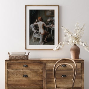 May include: A framed painting of a woman sitting in a chair in front of a vanity table. She is wearing a white robe and has her hair pulled back. She is looking at a mirror and holding a brush in her hand.