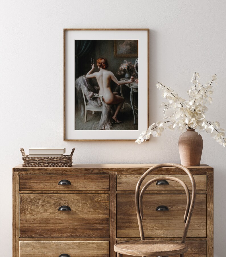 May include: A framed painting of a woman sitting in a chair in front of a vanity table. She is wearing a white robe and has her hair pulled back. She is looking at a mirror and holding a brush in her hand.