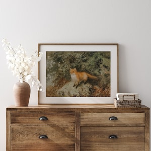 May include: A framed print of a red fox standing on a rock in a forest setting. The fox is looking to the left of the image. The print is in a brown wooden frame.