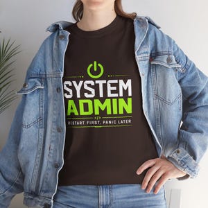 Könnte beinhalten: Ein braunes T-Shirt mit den Worten "SYSTEM ADMIN" in Weiß und Grün und dem Satz "RESTART FIRST, PANIC LATER". Das Shirt hat oben ein Stromsymbol. Die Person trägt eine Jeansjacke.