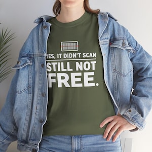 Op de afbeelding: Olijfgroen t-shirt met de tekst "YES, IT DIDN'T SCAN STILL NOT FREE." Een barcode-afbeelding staat boven de tekst. De persoon draagt een blauw spijkerjasje en een blauwe jeans.