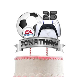 May include: A cake topper featuring a football, a video game controller, and a banner with the text "JONATHAN" and the number "25". The football has the EA Sports logo. The video game controller is white with black buttons. The banner is silver with black text.