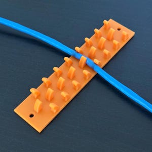 Könnte beinhalten: Ein orangefarbener Kabelorganizer aus Kunststoff mit mehreren kleinen, abgerundeten Vorsprüngen. Ein blaues Kabel ist über dem Organizer positioniert. Der Organizer hat zwei kleine Löcher, eines an jedem Ende. Der Hintergrund ist eine dunkle Oberfläche.