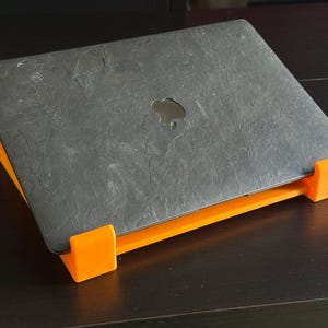 May include: A grey laptop computer with the Apple logo is on an orange laptop stand. The stand angles the laptop for better ergonomics. The stand is made of plastic. The laptop stand is approximately 30 cm long.