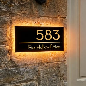 Peut inclure: Panneau d'adresse noir éclairé avec des chiffres et du texte dorés. Le panneau indique "583 Fox Hollow Drive" et est monté sur un mur en pierre. Le panneau est éclairé d'une lueur chaleureuse et accueillante.