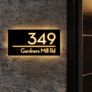May include: Black illuminated address sign with the number 349 and the street name Gardners Mill Rd in a modern font. The numbers and text are lit with a warm, golden glow against a black background. The sign is mounted on a textured, gray brick wall.