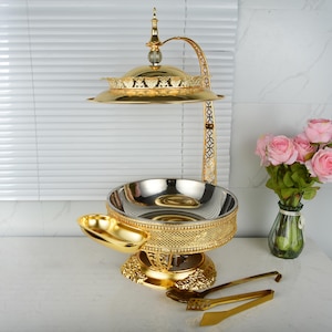 May include: A gold-colored, two-tiered serving stand with a decorative design. The top tier is a round, covered dish. The bottom tier is a large bowl with a silver interior. A small, gold-colored dish and serving tongs are also present.