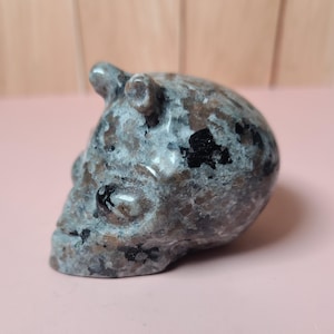 May include: A carved stone skull sculpture with a speckled pattern in shades of grey, brown, and black. The skull has two small horn-like protrusions on top. The sculpture is set against a pink background.