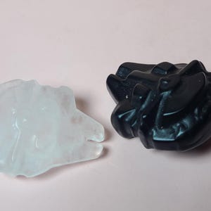May include: Two carved stone sculptures. One is translucent white, the other is black. Both are abstract, organic shapes. The white sculpture has a smooth, polished surface. The black sculpture has a textured, matte finish. The background is a solid light pink.