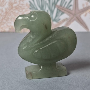 Pode incluir: Estatueta de pedra esculpida verde claro de um pássaro Dodo. O pássaro é retratado em posição sentada com um corpo arredondado, um bico curvo e uma pequena base quadrada. A estatueta tem um acabamento liso e polido.