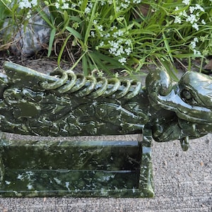 Puede incluir: Una escultura de dragón de jade tallado en tonos verdes, apoyada sobre un soporte a juego. El dragón presenta detalles intrincados, incluyendo escamas y una columna vertebral enrollada. La escultura se exhibe al aire libre.
