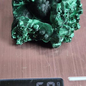 以下が含まれることがあります： 深いフォレストグリーンの色合いと質感のある結晶面を持つ、原石のマラカイト標本。鉱物の塊はデジタルスケールの上に置かれており、黒いデジタル画面には69グラムという重量がはっきりと表示されています。