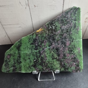 May include: A flat, triangular slab of zoisite mineral featuring a vibrant green base colour with prominent black hornblende inclusions and patches of deep magenta ruby. The stone is displayed upright on a clear, small, two-pronged metal stand.