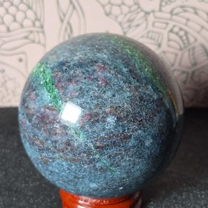 May include: A polished gemstone sphere with a mix of blue, green, and red hues. The sphere is resting on a small, round, reddish-brown wooden stand. The stone's surface has a textured appearance.