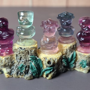 May include: Five colorful, carved figurines of people, each with a unique design and color, are displayed on a decorative base. The figurines are shades of purple, green, and pink. The base is a tree stump with green leaves.