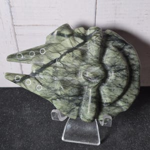 Puede incluir: Una escultura de piedra tallada con forma de nave espacial, con un cuerpo circular y dos puntas hacia adelante. El material es de color verde salvia jaspeado con vetas gris oscuro y pequeñas hendiduras circulares en la parte frontal.
