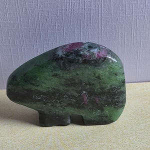 May include: A polished stone carving in the shape of a bear, featuring a deep green base colour with dark grey mottling and distinct, vibrant magenta-pink inclusions scattered across the upper portion of the smooth, rounded mineral surface.