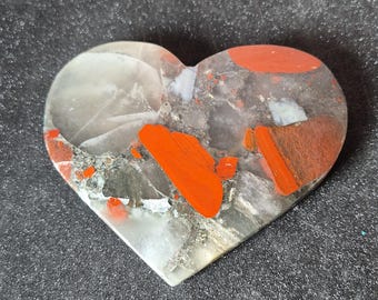 May include: A heart-shaped stone with a polished surface. The stone features a mix of gray, white, and red-orange colors. The red-orange sections are irregularly shaped and add a striking contrast to the other colors. The stone is smooth and appears to be a decorative item.