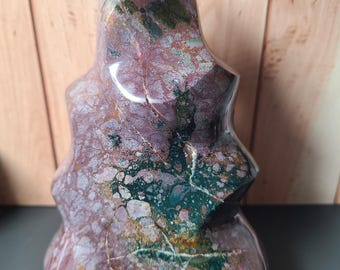 May include: A polished, freeform gemstone sculpture. The stone features a mix of colors, including deep burgundy, forest green, and cream. The sculpture has a textured surface and an organic shape, with a rounded base and a pointed top.