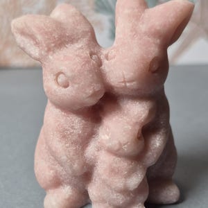 May include: A pink stone sculpture of three rabbits, two larger ones embracing a smaller one. The bunnies have detailed features, including carved eyes and noses. The sculpture is a light pink color and has a textured surface.
