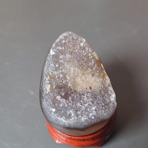 May include: A geode crystal with a purple and white interior, mounted on a small, round, reddish-orange wooden stand. The crystal has a rough, textured surface and a rounded shape. The stand provides a stable base for display.