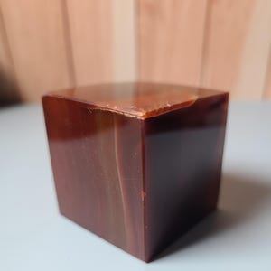 Puede incluir: Un cubo pulido de color marrón rojizo con una superficie superior ligeramente irregular. El cubo tiene un acabado liso y reflectante, y el color varía en tono en todo el material. El cubo es una forma geométrica sólida.