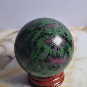 Può includere: Una sfera lucida di zoisite con cristalli di rubino incastonati, caratterizzata da un motivo maculato di verde bosco profondo, nero e magenta vibrante. La pietra poggia su un piccolo supporto circolare in legno color marrone rossastro.
