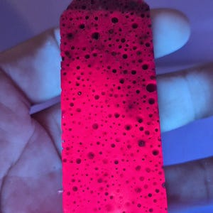 May include: A bright neon pink, porous, rectangular-shaped object with a pointed top, featuring a dense pattern of small, dark, circular indentations across its entire surface, illuminated by a vibrant light source from below.