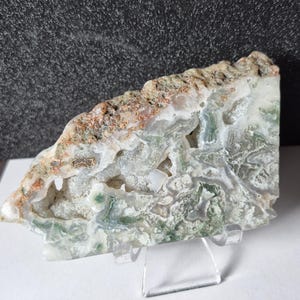 Puede incluir: Una piedra de ágata pulida, de forma triangular, que muestra una mezcla de colores blancos, verdes y marrones. La estructura cristalina de la piedra presenta cavidades visibles y se exhibe sobre un soporte transparente.