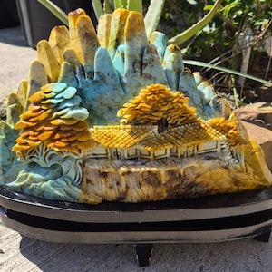 May include: A carved stone sculpture featuring a mountain landscape with a house and trees. The stone displays shades of blue, yellow, and brown. The sculpture is mounted on a smooth, dark, oval-shaped wooden base with small feet.