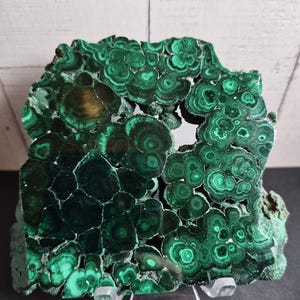 Könnte beinhalten: Ein poliertes Malachit-Exemplar mit tiefgrünen, konzentrischen kreisförmigen Mustern und Bänderungen. Die Mineralplatte weist eine natürliche, unregelmäßige Form mit einer zentralen Öffnung auf und steht auf einem transparenten Acrylständer.