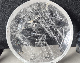 May include: A clear, round quartz crystal with internal fractures and inclusions. The crystal is displayed on a clear stand, showcasing its natural, translucent beauty. The quartz has a polished surface and a circular shape.