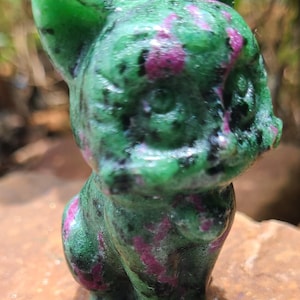 May include: A green and pink gemstone cat figurine. The cat is carved from a stone with a speckled pattern of green and magenta. The figurine has a smooth, polished surface and is posed in a seated position.