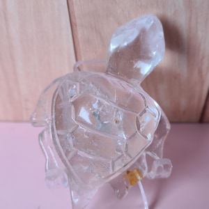 May include: A clear, glass turtle figurine with a detailed shell and head. The figurine has a small, amber-colored piece attached to its underside. The turtle is displayed on a light pink surface.