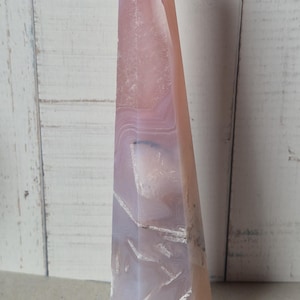 Peut inclure: Un obélisque en agate rose et blanche, de forme haute et pointue. La pierre présente une surface polie avec des motifs et des veines naturels. L'obélisque mesure environ 20 cm de haut.