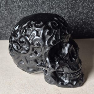 May include: A glossy black skull sculpture with intricate, swirling patterns on the cranium. The skull's eye sockets and nasal cavity are clearly defined, and the teeth are visible. The sculpture is a decorative item, possibly for Halloween or gothic decor.