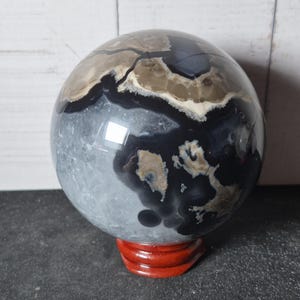 May include: A polished spherical stone featuring a complex pattern of deep black, charcoal gray, and creamy beige bands with natural inclusions. The sphere is supported by a small, circular, reddish-brown wooden stand with a smooth, glossy finish.