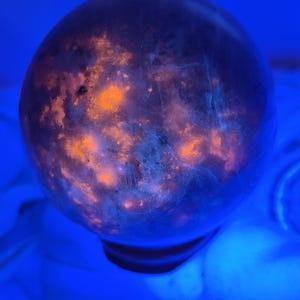 Puede incluir: Una esfera de piedra que muestra una superficie oscura y translúcida con inclusiones internas que fluorescen en naranja brillante y azul intenso bajo luz ultravioleta, creando una apariencia moteada similar a una galaxia en su forma.