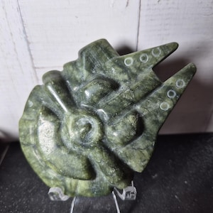 Può includere: Una scultura in pietra verde intagliata a forma di astronave con un corpo centrale circolare, un abitacolo sporgente e due punte rivolte in avanti. La superficie presenta una trama screziata con venature di colore verde scuro e verde chiaro.