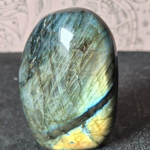 May include: A polished, oval-shaped labradorite stone. The stone displays a mix of colors, including blues, greens, and yellows, with visible internal fractures. The surface reflects light, highlighting the stone's iridescent qualities. The overall shape is smooth and rounded.