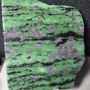 Pode incluir: Uma placa polida de zoisite com rubi. A pedra apresenta uma cor de base verde vibrante, acentuada por listras pretas e manchas de cristais de rubi rosa-avermelhados. A forma irregular e a superfície polida realçam a beleza natural do mineral.