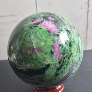 May include: A polished sphere of zoisite with inclusions of vibrant magenta ruby and dark black hornblende. The stone features a smooth, reflective surface with natural mineral patterns and sits atop a small, circular, dark brown wooden stand.