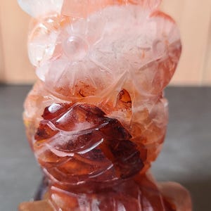 May include: A carved owl figurine made of translucent, reddish-orange crystal. The owl has detailed feather and facial features. The crystal has inclusions and colour variations. The owl is a decorative item.